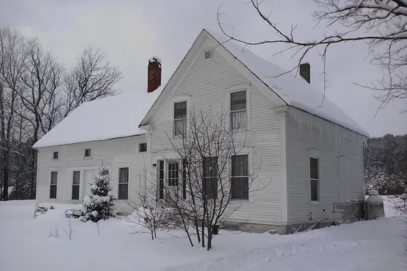 White Vermont farmhouse with two brick chimneys sitting in deep snow, bare maple trees standing beside it